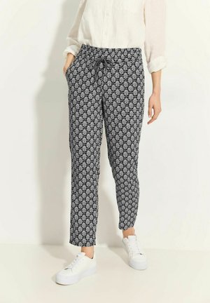 Frau trägt schwarz-weiß gemusterte, mit Kordelzug versehene Hosen mit Blumenmuster, weißes Langarmshirt und weiße Turnschuhe, steht mit den Händen in den Taschen.