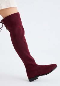 Mujer con una bota de ante burdeos hasta la rodilla, con detalle de cordones en el lateral y tacón bajo negro, sobre un fondo blanco.