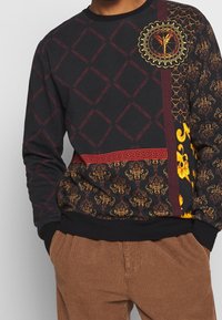 Sort sweatshirt med blandede mønstre: rød tekst, gyldne blomsterdesign og geometriske detaljer; ribbede manchetter og kant.