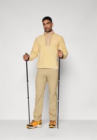 Gul fleecegenser med halvlås og frontlomme, kombinert med beige bukser og oransje tursko, ledsaget av vandrestaver.