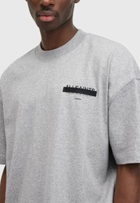 Camiseta de manga corta gris hecha de una tela suave, con un diseño frontal que presenta "ALL SAINTS" y un texto superpuesto en negro, estilo cuello redondo.