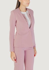 Donna che indossa un blazer su misura rosa e pantaloni abbinati su una maglietta bianca con decorazioni di perle, in piedi contro uno sfondo uniforme.