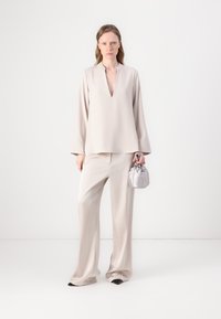 Tunica di un beige chiaro con scollatura a V profonda, abbinata a pantaloni larghi coordinati. Tiene una piccola borsa a tracolla argentata. Tessuto dalla consistenza liscia.
