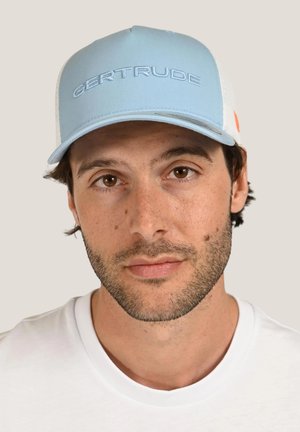 Gros plan d’un homme avec une légère barbe, portant une casquette bleu clair brodée de « GERTRUDE » et un t-shirt blanc à col rond.