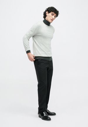 Lichtgrijze trui met zwarte accenten, ronde halslijn en lange mouwen, gecombineerd met donkere broek en zwarte schoenen. Vlakke textuur, moderne silhouet.