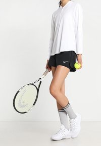 Camiseta blanca de manga larga con cremallera, pantalones cortos negros con el logo de Nike, calcetines grises, zapatillas de tenis blancas, sosteniendo una raqueta de tenis y una pelota.