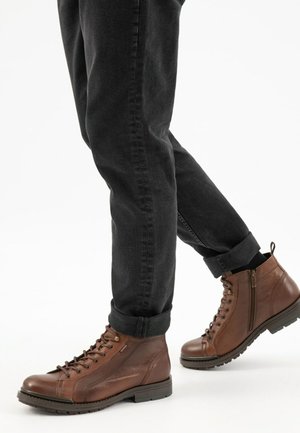 Bottines en cuir marron avec un laçage à l'avant, des côtés texturés et une semelle en caoutchouc. Un jean noir avec des ourlets retroussés visible à côté des bottines.
