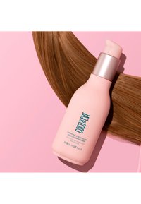 Fuktgivande och utredande leave-in balsam i en rosa flaska med en silverpump. Texten är i teal. Bruna hårstrån ligger bredvid flaskan.
