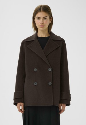 Femme portant un manteau en laine brun foncé à double boutonnage avec de larges revers et des manches retroussées, debout devant un fond clair uni.