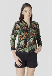 Jonge vrouw die een zwart jack met ritssluiting draagt met groene, oranje en roze tropische blaadjespatronen en zwarte korte broek, staand met handen in de zakken.