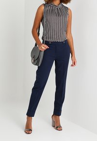 Ärmellose Bluse mit schwarz-weißen Punkten, navyfarbene Tailored Pants, graue Handtasche und Absatzsandalen, die einen eleganten, professionellen Look kreieren.