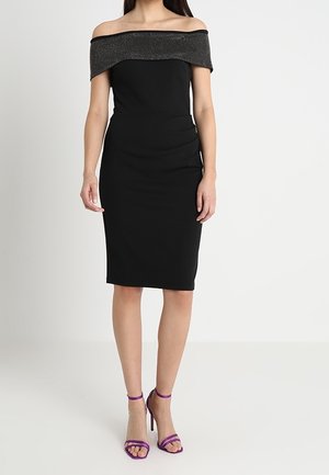 Vrouw draagt een zwarte nauwsluitende off-shoulder jurk met een sprankelende halslijn en paarse sandalen met hoge hakken, staand tegen een effen achtergrond.