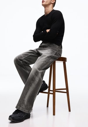 Mann trägt ein schwarzes langärmeliges Hemd, graue Jeans und schwarze Sneaker, sitzt auf einem hohen Holzstuhl vor weißem Hintergrund.