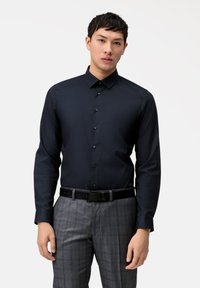 Marineblaue Langarmhemd mit Knopfleiste, spitzem Kragen und dunklen Knöpfen, kombiniert mit grauen karierten Hosen und einem schwarzen Gürtel.