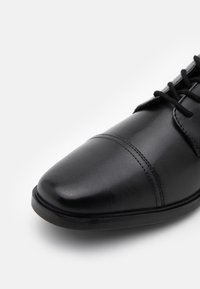 Black leather oxford shoe with a rounded toe, accent stitching, and a smooth finish. Features flat laces and a low-profile rubber sole.