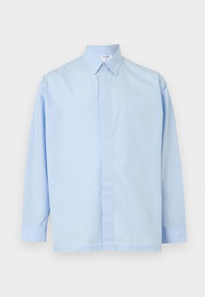 Homme aux cheveux bouclés portant une chemise boutonnée bleu clair oversize, un pantalon noir et des chaussures habillées noires, debout avec une main dans la poche.