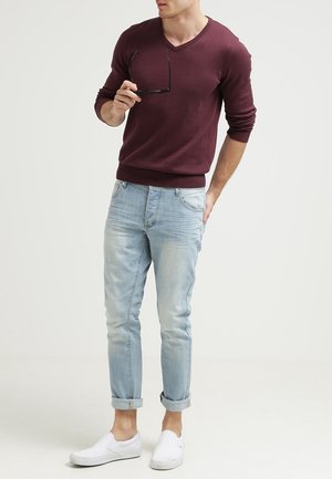 Mann in kastanienbraunem Langarmpullover, hellblauen hochgekrempelten Jeans und weißen Slip-On-Schuhen hält schwarze Sonnenbrille vor einfachem hellem Hintergrund.