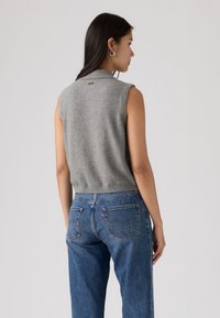 Γυναίκα με μακριά σκούρα μαλλιά που φορά ασημένια σκουλαρίκια κρίκους, γκρι αμάνικο πλεκτό γιλέκο και μπλε τζιν, στραμμένη πλευρικά σε μια απλή φόντο.