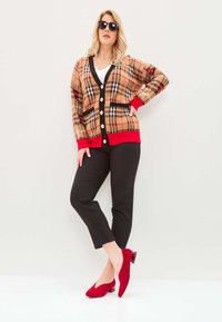 Beige Karoblazer mit roten Akzenten, schwarzem Besatz und großen Knöpfen. Schwarze Hose und rote Wildlederabsätze runden das Outfit ab.