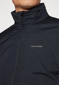 Schwarze leichte Jacke mit hohem Kragen, Frontreißverschluss und kleinem reflektierendem Logo auf der linken Seite. Strukturierter Stoff, elegantes Design.