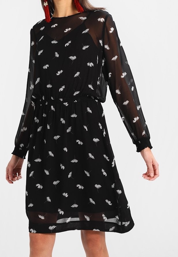 Vestido negro de longitud a la rodilla hecho de tela transparente, con un patrón floral en blanco. Mangas largas y cintura elástica, con un dobladillo ligeramente en capas.