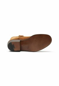 Zapato de ante marrón con suela plana, que presenta una punta redondeada y un lazo decorativo. La suela tiene un acabado texturizado y un logo en relieve.
