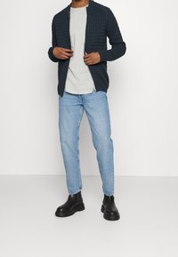 Man i svart texturerad zip-up cardigan, ljusgrå t-shirt, ljusblå jeans och svarta läderstövlar står mot vit bakgrund.