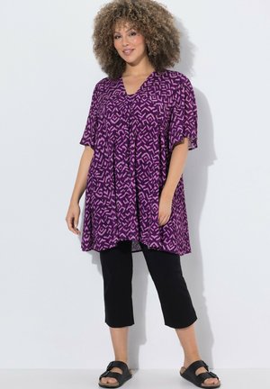 Frau mit lockigem Haar, die eine lila gemusterte Tunika, schwarze kurze Hosen und schwarze Sandalen trägt, steht vor einem einfarbigen weißen Hintergrund.