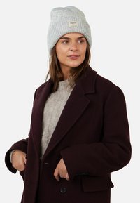 Berretto in maglia grigia con texture a coste e un'etichetta rettangolare. Indossato con un maglione grigio chiaro e un cappotto bordeaux scuro.
