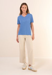 Modrá v-neck tričko z lehkého materiálu, spárované s krémovými širokými kalhotami a bílými teniskami s modrými akcenty. Viditelná měkká textura.
