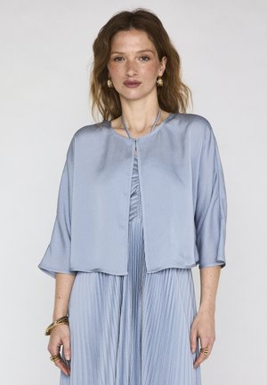 Donna con capelli castani ondulati che indossa un abito plissettato azzurro chiaro e una giacca ampia abbinata, accessoriata con orecchini e braccialetti dorati.