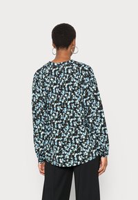 Blusa preta com padrão floral azul, mangas longas com punhos elásticos, decote redondo e um corte relaxado. A vista traseira mostra um leve detalhe de franzido.