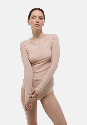 Femme aux cheveux bruns portant un haut beige ajusté à manches longues et une lingerie assortie, ajustant sa manche gauche sur un fond blanc uni.