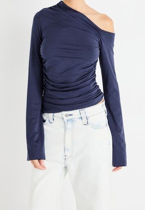 Person trägt ein marineblaues schulterfreies Langarmoberteil mit gerafften Details und hellblaue Jeans mit hoher Taille.