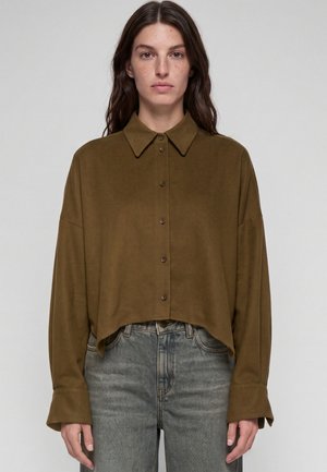 Frau trägt eine locker sitzende olivgrüne Bluse mit Knöpfen, langen Ärmeln und hoch taillierte ausgewaschene graue Jeans, stehend vor einem schlichten Hintergrund.