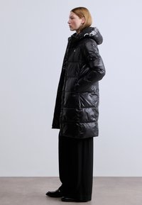 Μαύρο, φουσκωτό παρκά με γυαλιστερό φινίρισμα, με ευρύχωρη κουκούλα και πλαϊνές τσέπες. Ο σχεδιασμός περιλαμβάνει οριζόντια ραμμένα πάνελ.