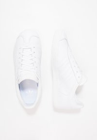 White sneakers with smooth leather material, flat laces, and rubber sole. Features three striped accents on the side and a rounded toe design.