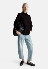 Donna che indossa una camicia nera con bottoni, jeans strappati blu chiaro, mocassini neri, mentre tiene una grande borsa nera con catena dorata su sfondo bianco.