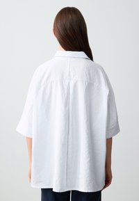 Camicia bianca oversize con colletto, maniche corte, tessuto texturizzato e cucitura a yoke sul retro, indossata sopra pantaloni blu navy.