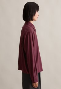 Burgundy blouse with a classic collar, long sleeves with button cuffs, and a loose fit, made of a smooth, lightweight fabric.