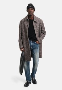 Brun herringbone-kappa, svart tröja och blå jeans. Bär en svart tote-väska. Svart beanie och glänsande svarta skor fulländar looken.