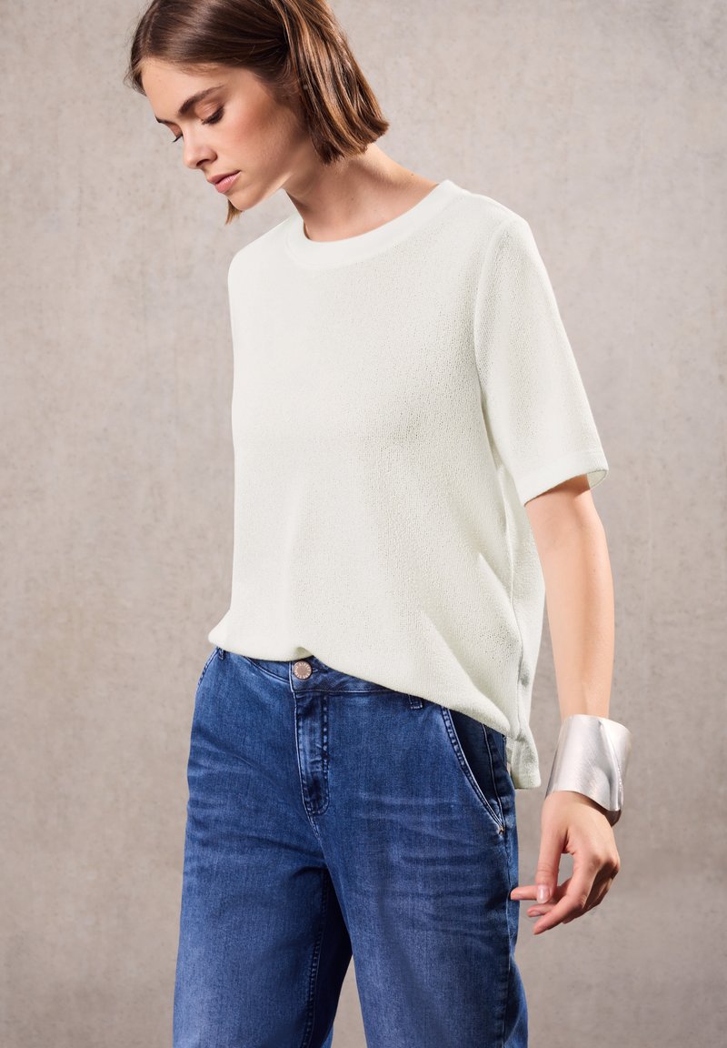 Wit, gestructureerd shirt met korte mouwen gecombineerd met blauwe spijkerbroeken. Opvallende zilveren armband om de pols. Eenvoudig, casual ontwerp.