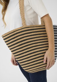 Borsa a tracolla a righe larghe realizzata in materiali naturali intrecciati, nei colori beige e nero, con manico intrecciato e bordo sfrangiato.