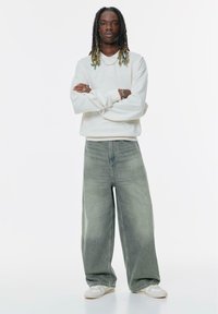 Sweatshirt alb cu mânecă lungă și guler rotund, blugi evazați de un verde estompat, accesorii argintii și adidași albi. Design simplu, material texturat.