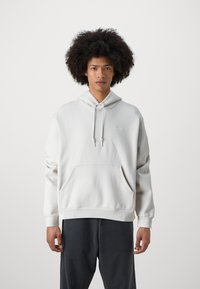 Mladý muž s kudrnatými vlasy, oblečený v bílém mikině Nike a tmavých kalhotách, stojící proti jednoduchému světlému pozadí.