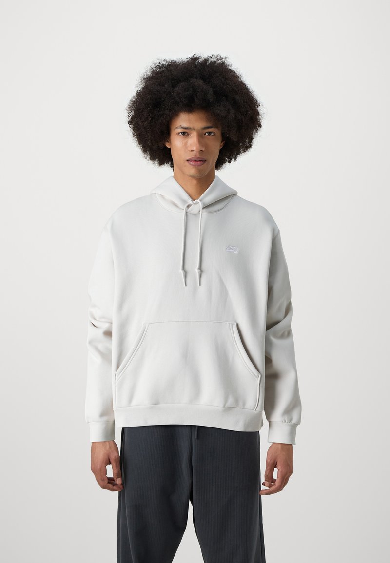 Mladý muž s kudrnatými vlasy, oblečený v bílém mikině Nike a tmavých kalhotách, stojící proti jednoduchému světlému pozadí.