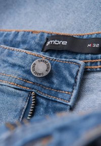 Farkut vaaleansinisinä, joissa on oranssit tikkaukset, pyöreä metallinappi, jossa on teksti "OMBRE DENIM", ja musta etiketti.