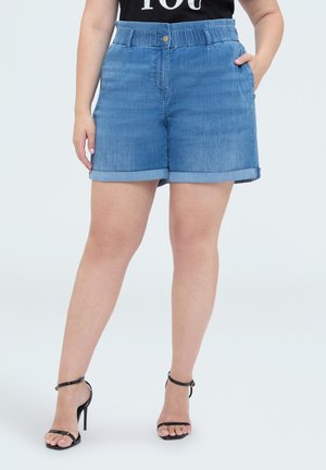 Persoon die blauwe denimshorts en zwarte sandalen met hoge hakken draagt, staand met één hand in de zak tegen een effen achtergrond.