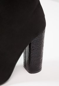 Zapato de tacón alto negro con un tacón bloque negro texturizado y brillante, y parte superior de ante suave. El tacón tiene una base cuadrada para mayor estabilidad.