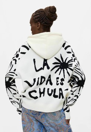 Kvinde med flettet hår bærer en hvid hættetrøje med sorte abstrakte designs og teksten "LA VIDA ES CHULA" i sort på ryggen.